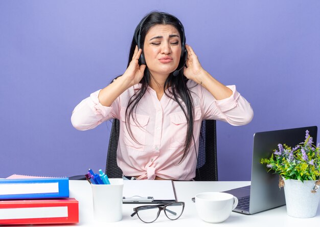 Young beautiful woman in casual clothes wearing headset with microphone looking annoyed suffering from noise sitting at the table with laptop over blue wall working in office