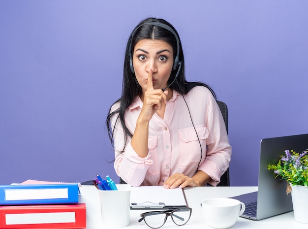 Young beautiful woman in casual clothes wearing headset with microphone making silence gesture with finger on lips worried sitting at the table with laptop over blue wall working in office