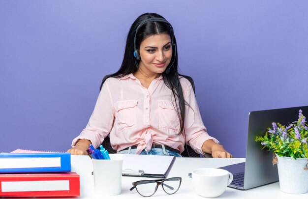 Young beautiful woman in casual clothes wearing headset with microphone sitting at the table with laptop looking at screen smiling confident over blue wall working in office