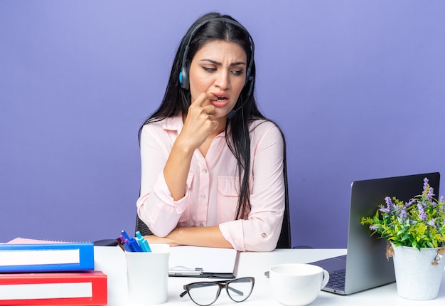 Young beautiful woman in casual clothes with headphones and microphone looking worried biting nails sitting at the table with laptop on blue