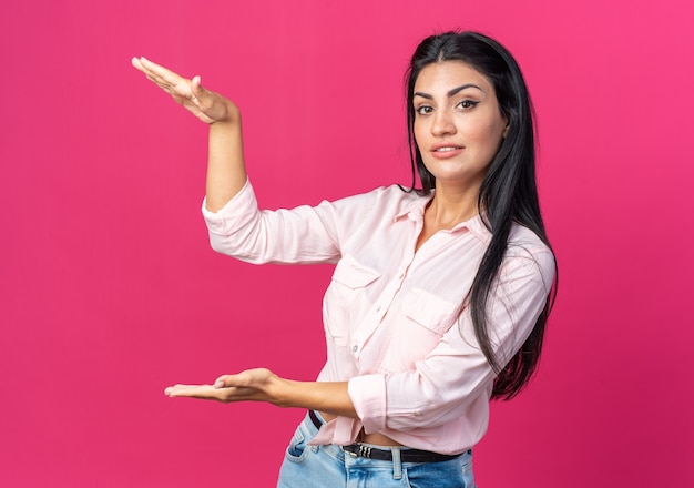 Молодая красивая женщина в повседневной одежде с серьезным лицом показывает жест большого размера руками, концепция символа измерения, стоящая над розовой стеной