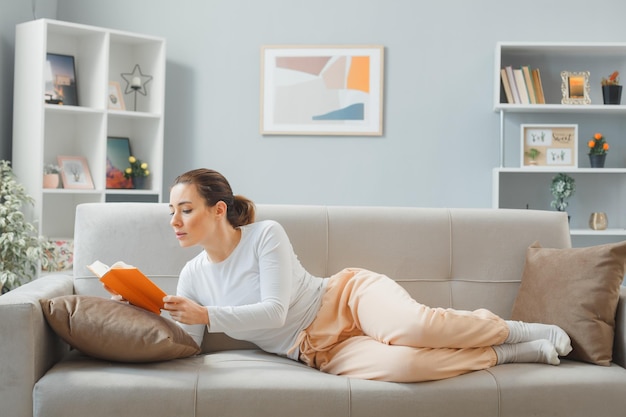 Free photo young beautiful woman in casual clothing laying on a couch at home interior reading a book looking confident happy and positive spending weekend at home