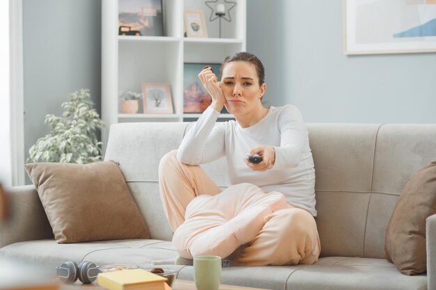 Young beautiful woman in casual clothing sitting on a couch at home interior eating cookie holding remote looking at camera confused and displeased spending weekend at home