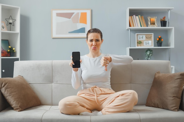 Young beautiful woman in casual clothing sitting on a couch at home interior showing smartphone and thumb down being displeased making wry mouth spending time at home