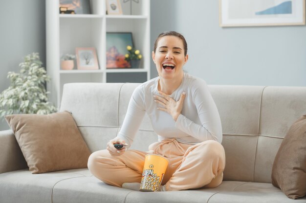 young beautiful woman in casual clothing sitting on a couch at home interior with bucket of popcorn holding remote looking at camera happy and joyful smiling broadly spending time at home