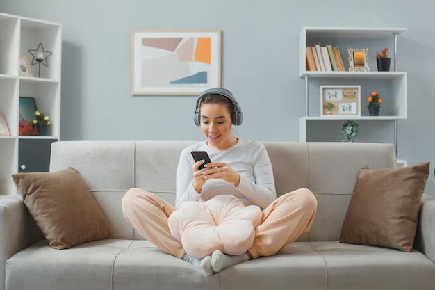 Молодая красивая женщина в повседневной одежде с наушниками сидит на диване в домашнем интерьере, держа смартфон, счастливая и удивленная, глядя на экран своего мобильного телефона, весело улыбаясь, расслабляясь