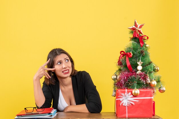 Young beautiful woman confused about something surprisedly sitting at a table near decorated Christmas tree at office on yellow 