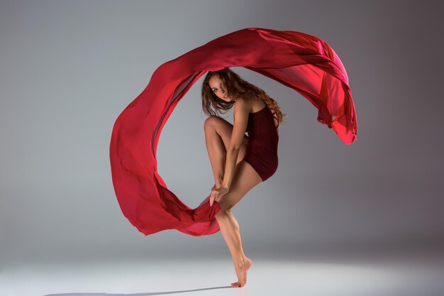 Молодая красивая танцовщица в красном купальнике позирует на светло-сером студийном фоне. Современный