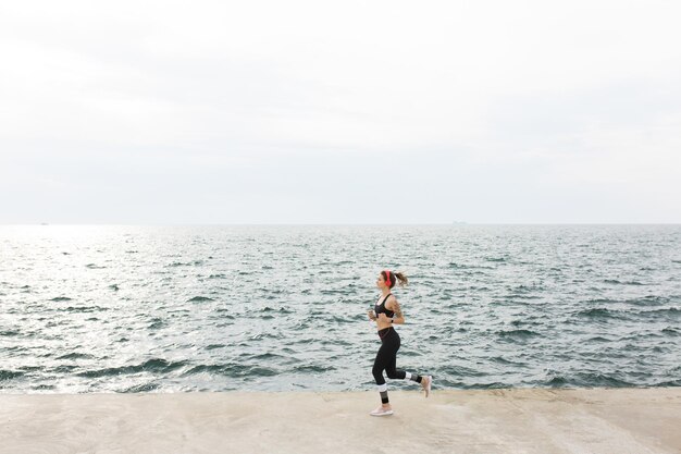 Young beautiful woman in dark gray sporty top and leggings with red headphones dreamily running with amazing sea view on background