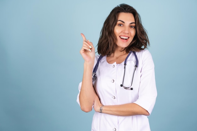 Young beautiful woman doctor in a lab coat on a blue background suddenly had an idea, points her finger up