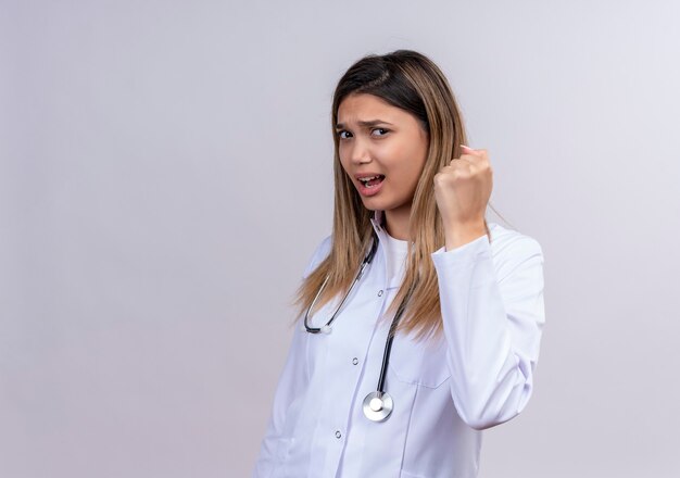 Молодая красивая женщина-врач в белом халате со стетоскопом, сжимающим кулак с сердитым лицом
