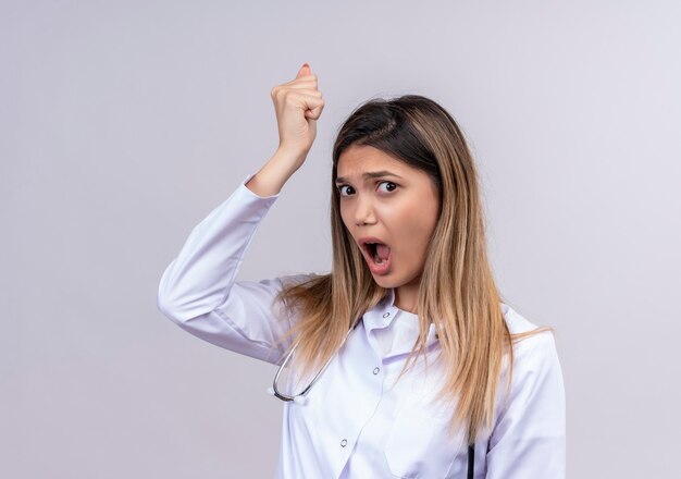 Young beautiful woman doctor wearing white coat with stethoscope clenching fist with angry face