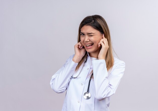 聴診器で白いコートを着た若い美しい女性医師は、大きな音のノイズのためにイライラした表情で指で耳を閉じます