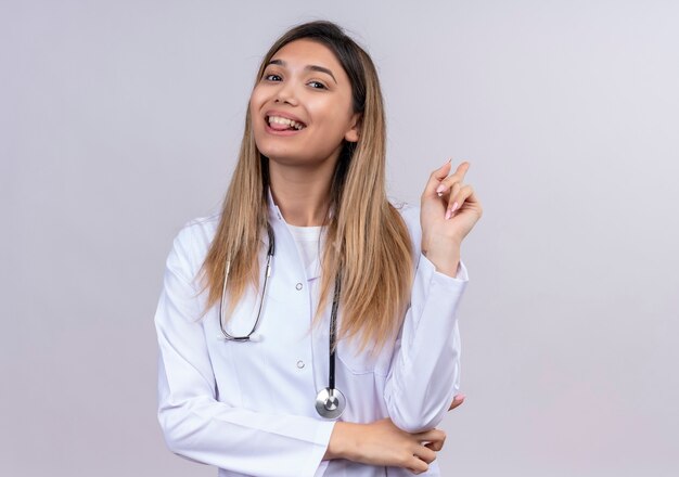 Молодая красивая женщина-врач в белом халате со стетоскопом, счастливая и позитивная, указывая назад с весело улыбаясь указательным пальцем