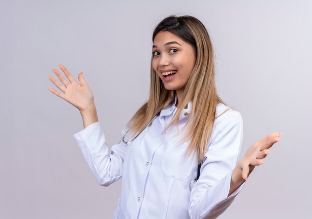 Young beautiful woman doctor wearing white coat with stethoscope happy and positive smiling wide opening hands making welcoming gesture