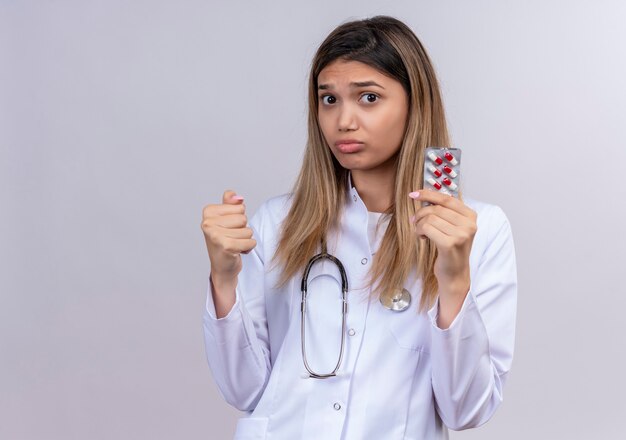Young beautiful woman doctor wearing white coat with stethoscope holding blister with pills clenching fist looking displeased