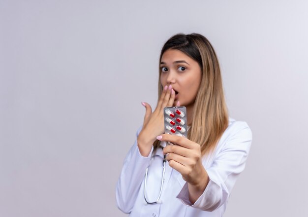 Young beautiful woman doctor wearing white coat with stethoscope holding blister with pills looking amazed surprised covering mouth with hand