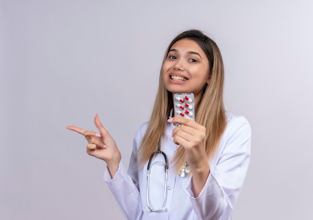 聴診器で白衣を着た若い美しい女性医師は、横に指で元気に笑顔の丸薬で水ぶくれを保持します