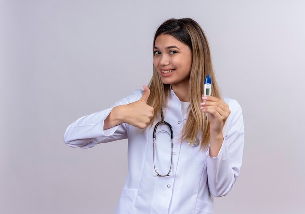 Молодая красивая женщина-врач в белом халате со стетоскопом держит цифровой термометр, улыбаясь счастливым лицом, показывает палец вверх