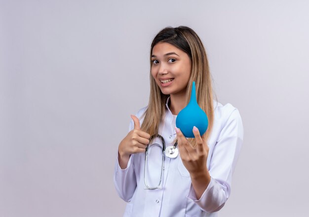 Young beautiful woman doctor wearing white coat with stethoscope holding an enema smiling showing thumbs up