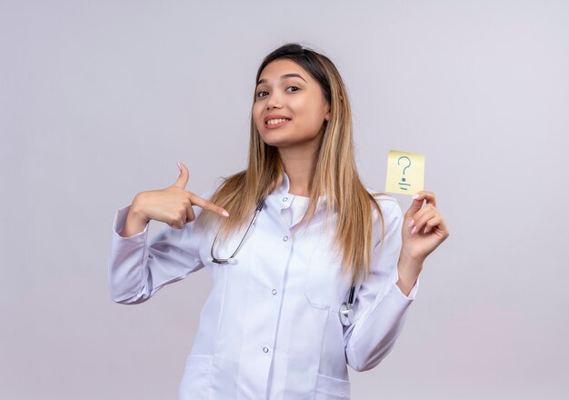 Молодая красивая женщина-врач в белом халате со стетоскопом держит бумагу для напоминания с вопросительным знаком, указывая указательным пальцем на нее положительно и счастливо