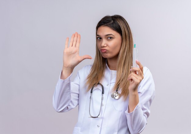 聴診器と注射器を保持している白いコートを着ている若い美しい女性医師は、一時停止の標識を作る開いた手で立って不快に見えます