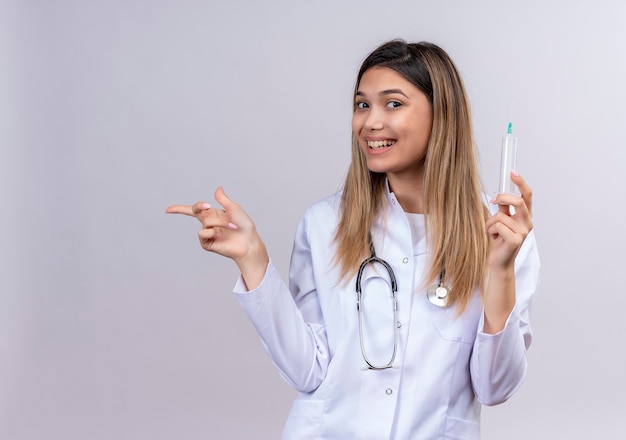 Молодая красивая женщина-врач в белом халате со стетоскопом держит шприц, весело улыбаясь, указывая указательным пальцем в сторону