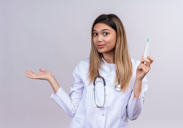 Молодая красивая женщина-врач в белом халате со стетоскопом, держащим шприц