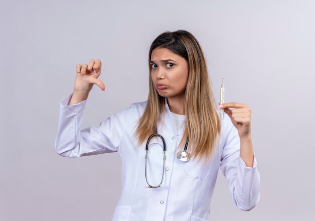 Young beautiful woman doctor wearing white coat with stethoscope holding thermometer looking with frowning face showing thumbs down