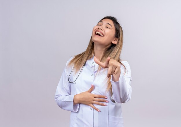 Young beautiful woman doctor wearing white coat with stethoscope joking with someone laughing pointing with index finger