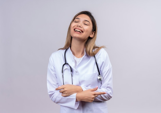 Young beautiful woman doctor wearing white coat with stethoscope laughing hugging her belly