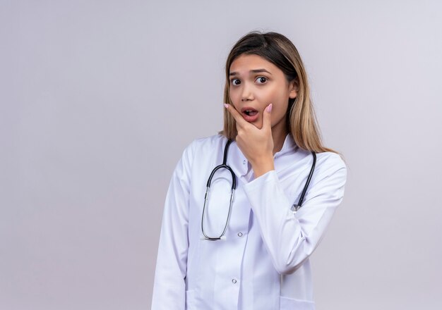 Young beautiful woman doctor wearing white coat with stethoscope looking amazed and surprised