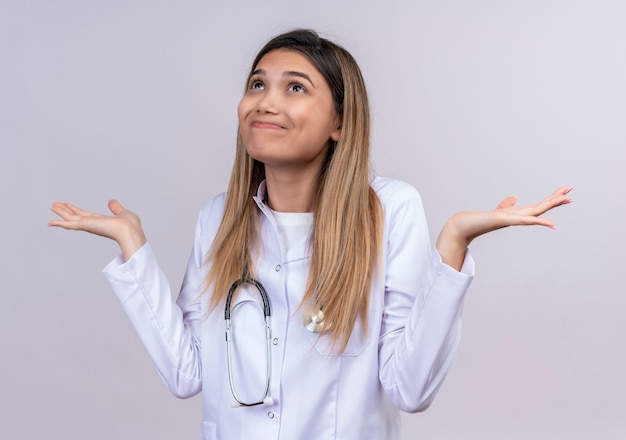 Giovane bella donna medico indossa camice bianco con lo stetoscopio cercando confuso e incerto scrollare le spalle non avendo risposta