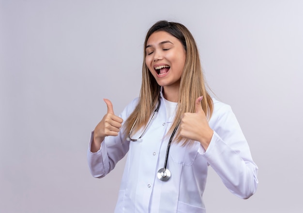 Young beautiful woman doctor wearing white coat with stethoscope looking happy and exited showing thumbs up