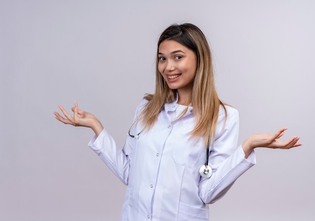 Молодая красивая женщина-врач в белом халате со стетоскопом выглядит позитивно и счастливо улыбается, разводя ладони в стороны