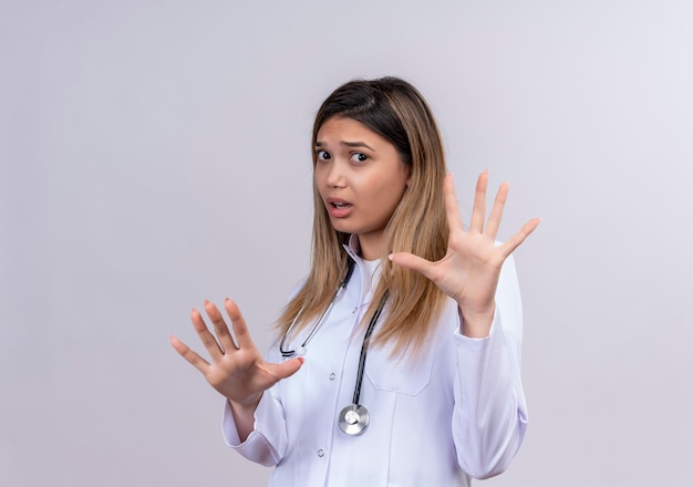 Young beautiful woman doctor wearing white coat with stethoscope looking scared making defense gesture raising open palms