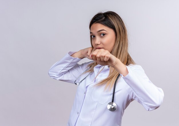 Young beautiful woman doctor wearing white coat with stethoscope looking with serious face posing like a boxer with clenched fists