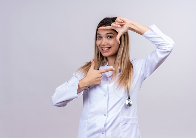 Young beautiful woman doctor wearing white coat with stethoscope making frame with fingers smiling looking through this frame