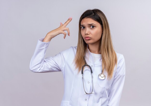 Young beautiful woman doctor wearing white coat with stethoscope making gun or pistol gesture near temple frowning face