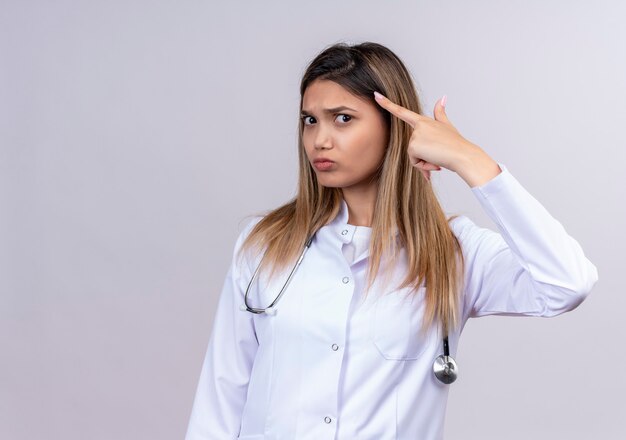 Young beautiful woman doctor wearing white coat with stethoscope pointing temple with finger concentrating hard on an idea with serious expression