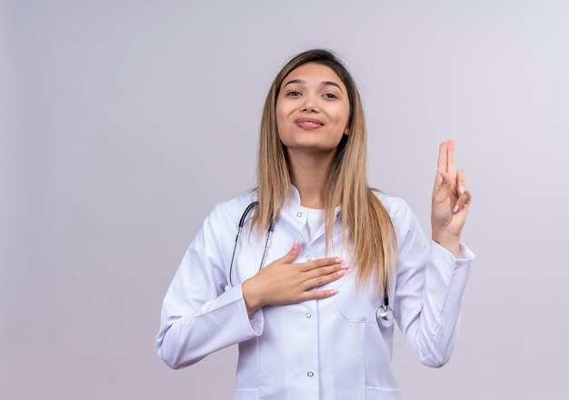 Young beautiful woman doctor wearing white coat with stethoscope raising hand to taking oath looking confident