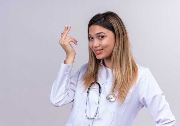 Young beautiful woman doctor wearing white coat with stethoscope smiling cheerfully gesturing with hand