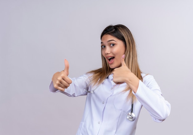 Молодая красивая женщина-врач в белом халате со стетоскопом, весело улыбаясь, показывает палец вверх