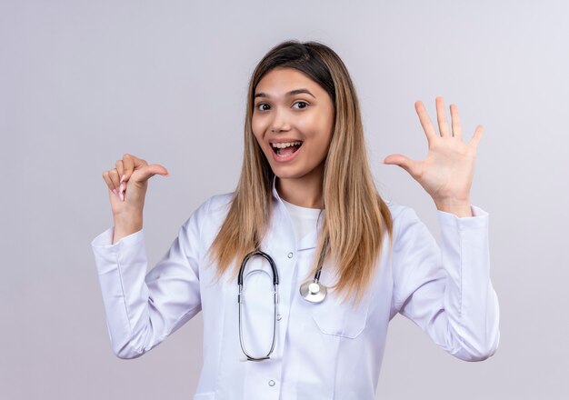 聴診器で白衣を着た若い美女医師が自信を持って見せて、指で上向きに笑顔6番