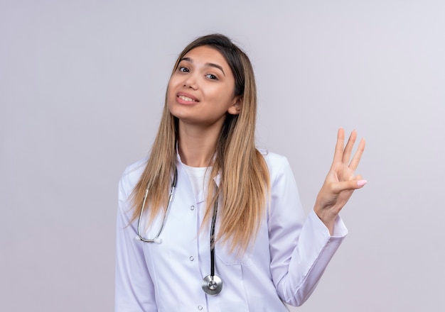 Молодая красивая женщина-врач в белом халате со стетоскопом, уверенно улыбаясь, показывает пальцами номер три