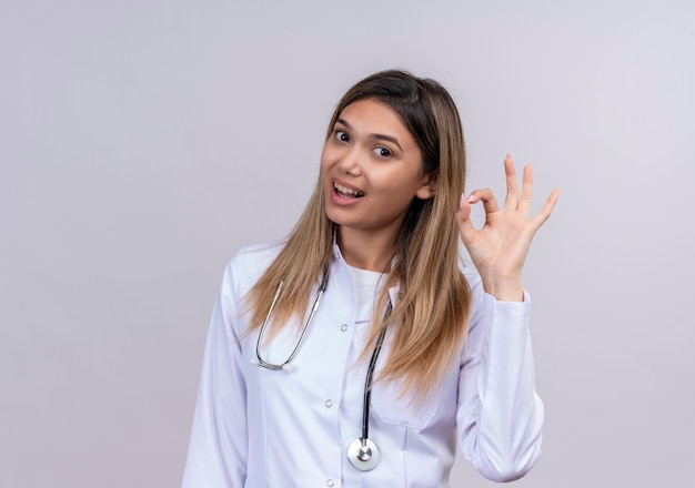 Giovane bella donna medico indossa camice bianco con lo stetoscopio sorridente amichevole facendo segno giusto