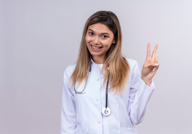 Young beautiful woman doctor wearing white coat with stethoscope smiling friendly showing victory sign