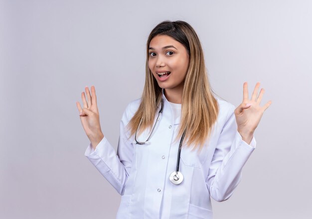 Young beautiful woman doctor wearing white coat with stethoscope standing with raised arms in surrender