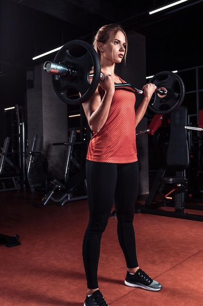 Giovane bella donna che fa esercizio con la barra in una palestra. ragazza atletica che fa allenamento in un centro fitness. su uno sfondo scuro la palestra. vestito con abiti sportivi.