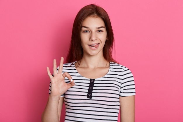 Молодая красивая женщина делает нормальный знак пальцами и показывает свой язык, с забавным выражением лица, позирует изолированно над розовой стеной.